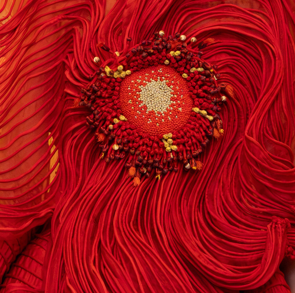 A model wearing a dramatic, voluminous red gown with sculptural layered ruffles and a large circular floral embellishment at the chest, posed in front of a warm-toned decorative mural.
