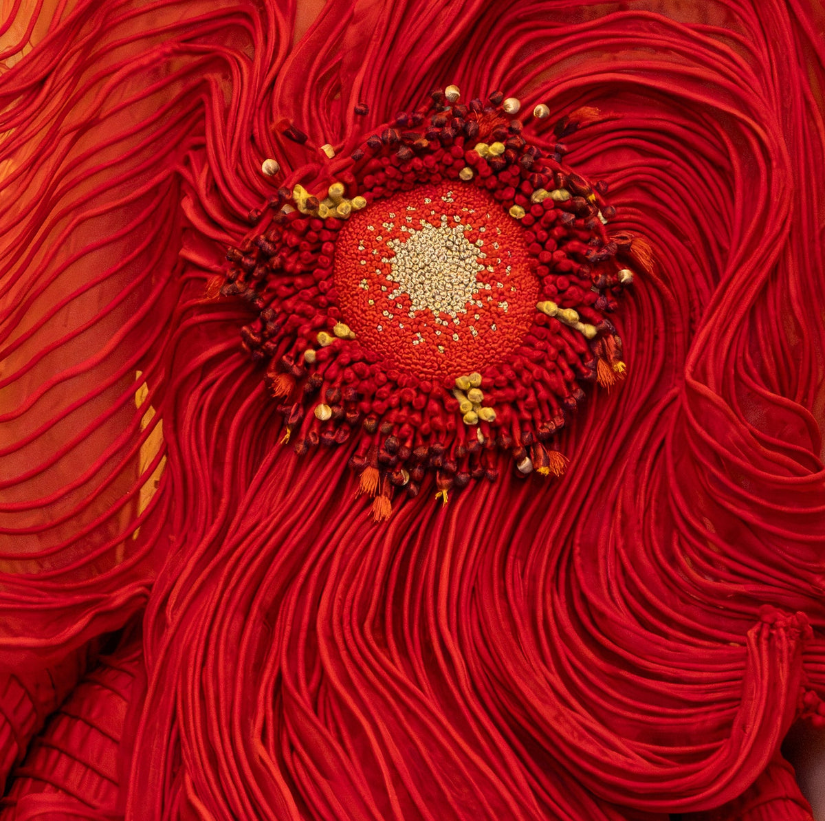 A model wearing a dramatic, voluminous red gown with sculptural layered ruffles and a large circular floral embellishment at the chest, posed in front of a warm-toned decorative mural.