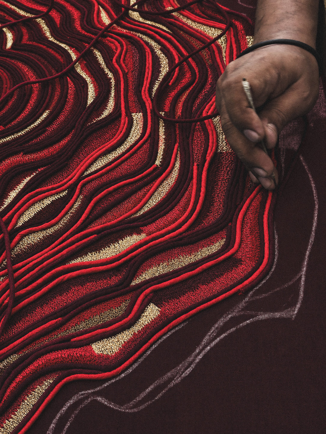 A close-up of a hand holding a needle and thread, working on a dark red fabric with layered wavy embroidery in red, black, and metallic gold.